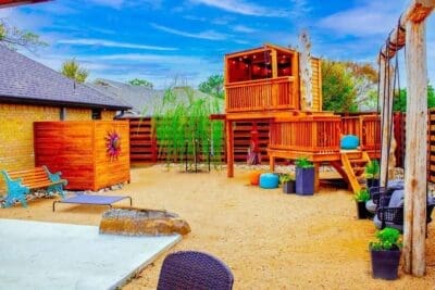 Backyard play area in Prestonwood Dallas, TX with decomposed granite, raised wooden playhouse, privacy fence, bench, boulder, and container plants