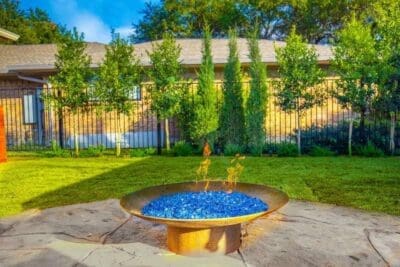 Steel fire bowl with blue fire glass in a Far North Dallas backyard with flagstone patio, lawn, privacy trees, and metal fencing