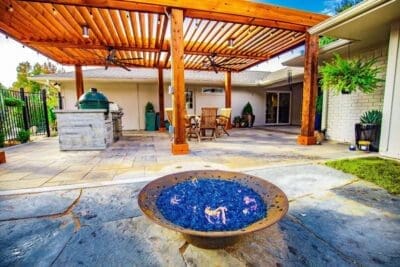 Steel fire bowl with blue fire glass in a Far North Dallas backyard with stone patio, wood patio cover, outdoor kitchen, and hanging plants