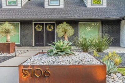 Corten steel address planter in Russwood Acres Dallas, TX with agave, yucca, river rock, and modern walkway near a painted brick home