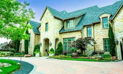 Front yard landscape design in North Dallas, TX with a paver driveway, curved stone border, decorative pots, layered shrubs, seasonal flowers, boulders, and landscape lighting.