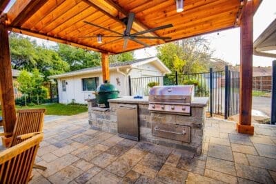 Built-in stone outdoor kitchen in Far North Dallas with grill, smoker, refrigerator, paver patio, wood patio cover, and ceiling fan