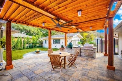 Covered outdoor living patio in Far North Dallas with wood pergola-style roof, outdoor kitchen, paver patio, dining area, fire feature, and lawn