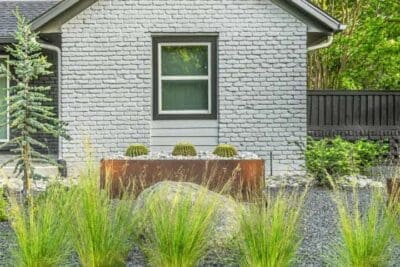 Side yard xeriscape planting in Russwood Acres Dallas, TX with Corten steel planter, ornamental grasses, gravel, boulders, and painted brick exterior