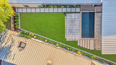 Overhead view of modern backyard landscape design in Northwood Heights, Dallas, TX with turf lawn, geometric pool, covered pergola, concrete walkway, gravel border, and perimeter plantings.