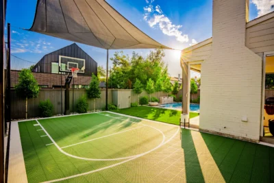 Custom backyard basketball court with overhead shade structure, turf lawn, pool area, and privacy planting in Midway Hollow Dallas, Texas.