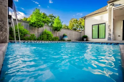 Modern backyard pool with raised water feature, privacy planting, and covered patio beside a white brick home in Midway Hollow Dallas, Texas.