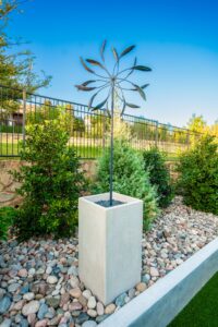 Decorative metal wind sculpture in a Hidden Knoll backyard planting bed with river rock, shrubs, black metal fencing, and artificial turf in Southlake, Texas