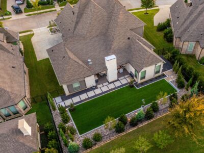 Aerial backyard landscape design in Hidden Knoll with artificial turf, square stepping stones, river rock beds, metal planters, and privacy plantings in Southlake, Texas