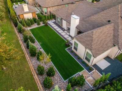 Aerial view of a Hidden Knoll backyard with artificial turf, river rock borders, square concrete pavers, shrubs, and a modern side yard layout in Southlake, Texas