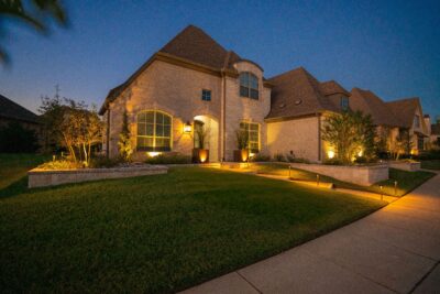 Nighttime front yard in Hidden Knoll, Southlake, Texas, with warm path lights, uplighting, stone raised beds, shrubs, metal planters, and a curved walkway