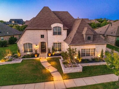 Evening view of a Southlake, Texas front yard with landscape lighting, stone raised beds, river rock, ornamental shrubs, and a lit walkway