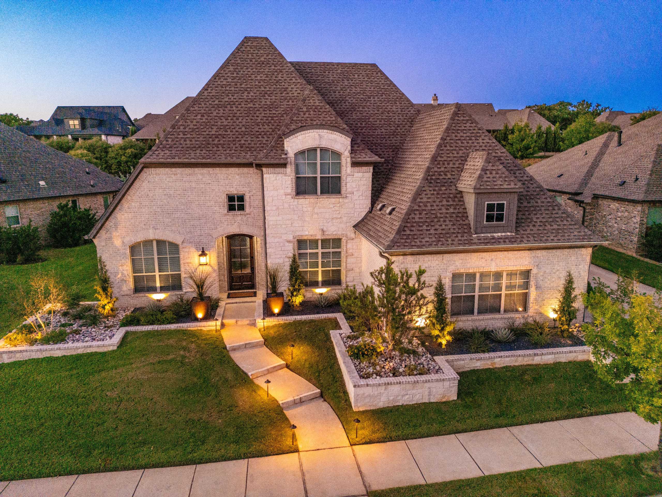 Evening view of a Southlake, Texas front yard with landscape lighting, stone raised beds, river rock, ornamental shrubs, and a lit walkway