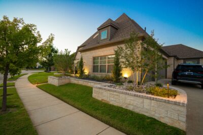 Front yard raised stone planting beds in Southlake, Texas, with warm landscape lighting, shrubs, trees, river rock, and a curved sidewalk
